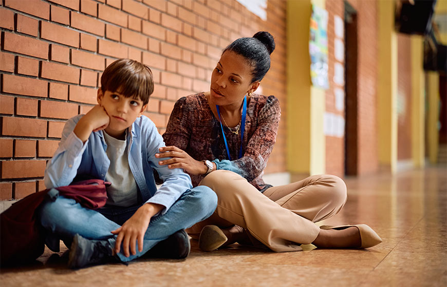 Photo of a teacher talking to a sad boy in the hallway of a school
